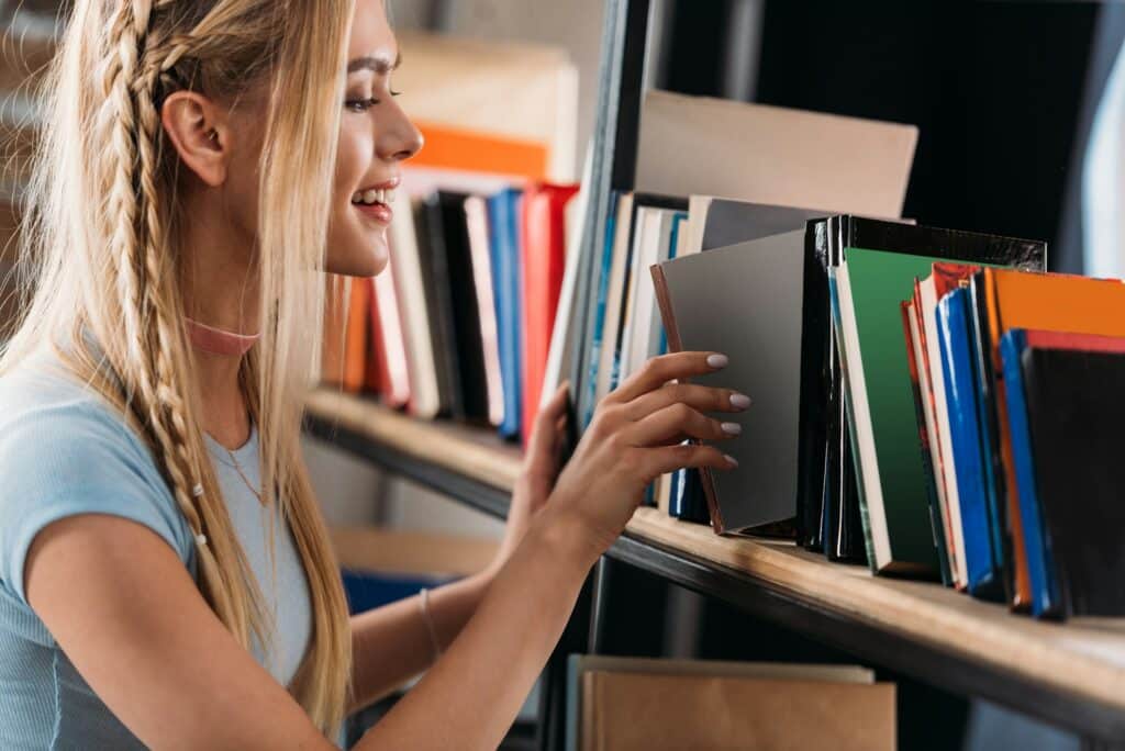 Smiling young woman choosing book on bookshelf in library